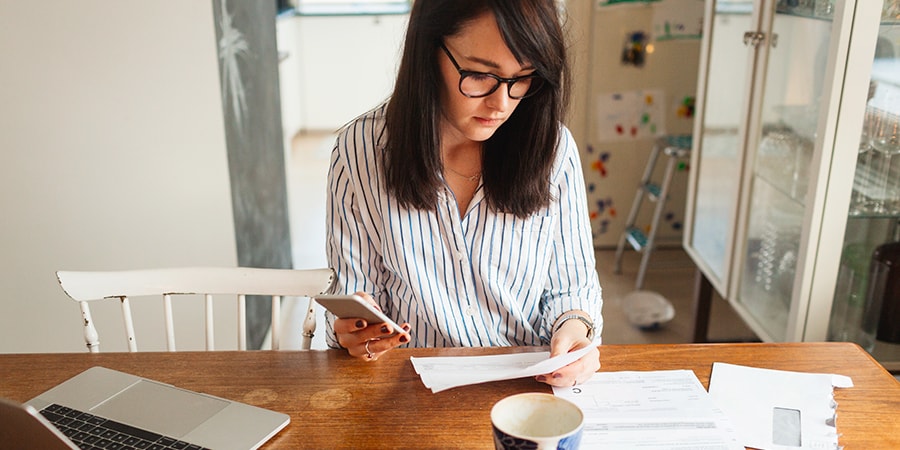 Dame sitter på kjøkkenbordet med mobil i hånda og leser dokumenter, til illustrasjon for dødsbo og arveoppgjør