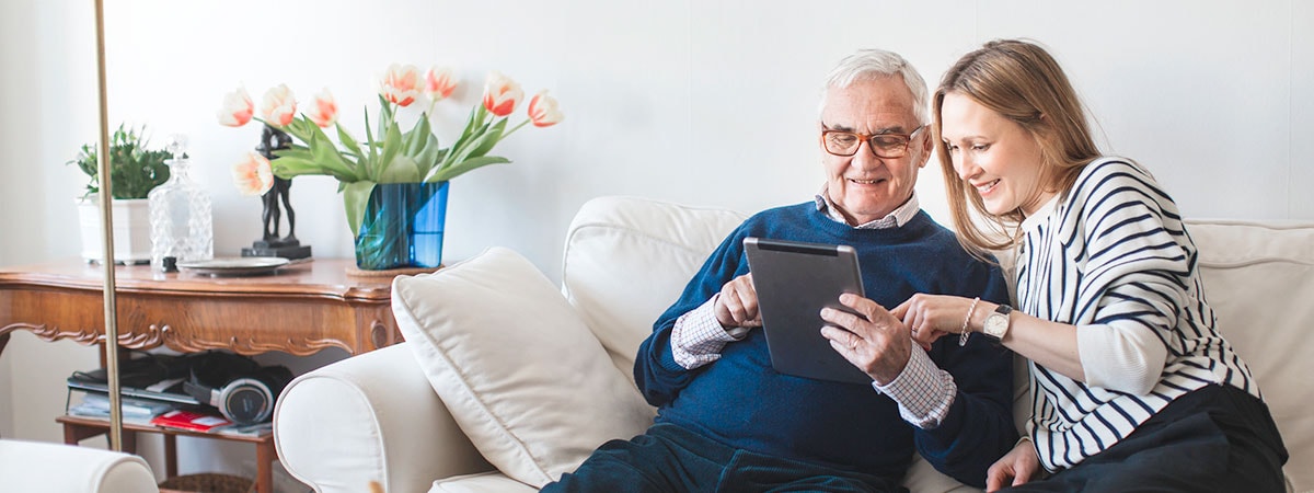 Eldre mann og ung kvinne sitter i en sofa med et nettbrett og skriver fremtidsfullmakt