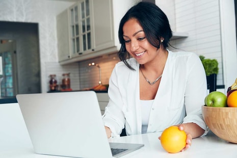 Dame sitter på kjøkkenet og jobber på PC mens hun holder en appelsin, til illustrasjon for brevgiro