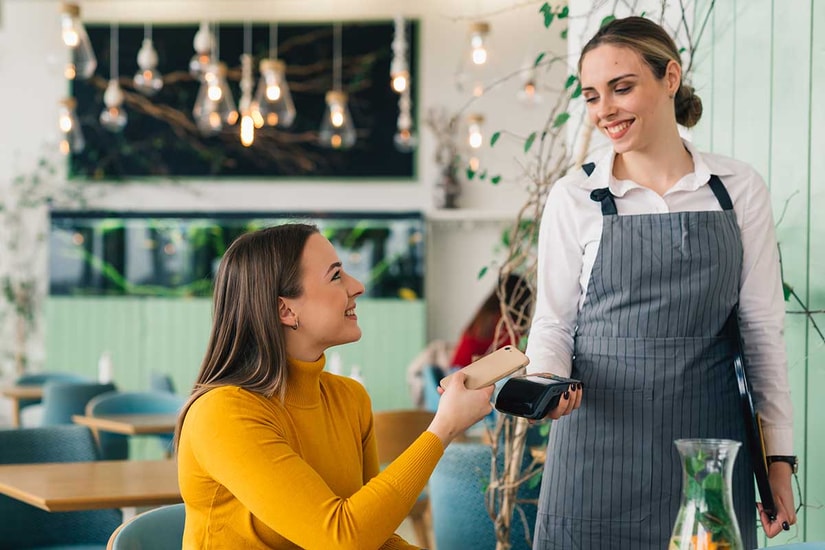 Dame sitter på kafé og betaler med mobiltelefonen til servitør