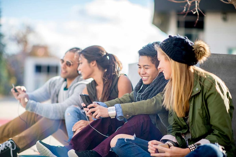 Ungdommer sitter på bakken og ser på mobilene sammen, til illustrasjon for bank for barn og ungdom
