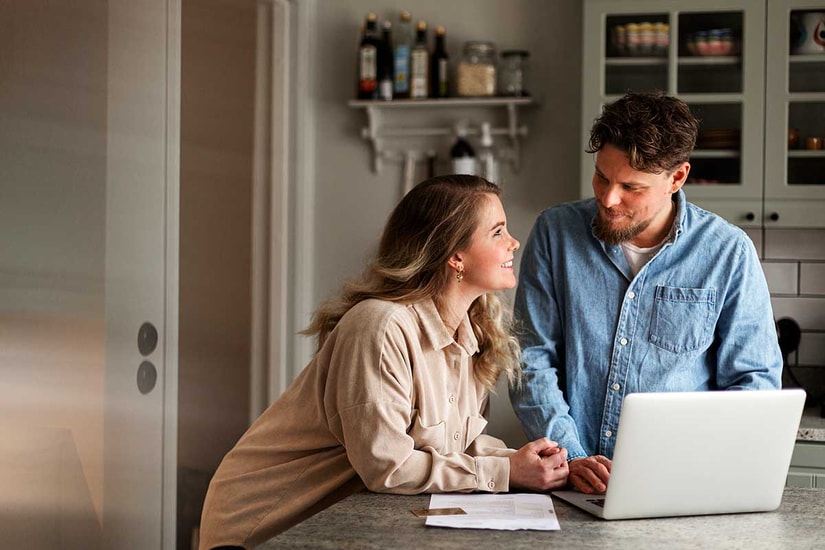 Kvinne og mann står på kjøkkenet og lener seg over kjøkkenbenken med en PC fremfor seg, til illustrasjon for annuitetslån vs serielån