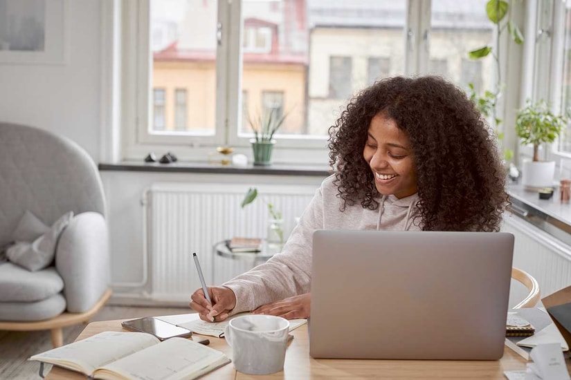 Dame sitter på spisebord og skriver notater med en PC fremfor seg, til illustrasjon for refinansiere forbruksgjeld