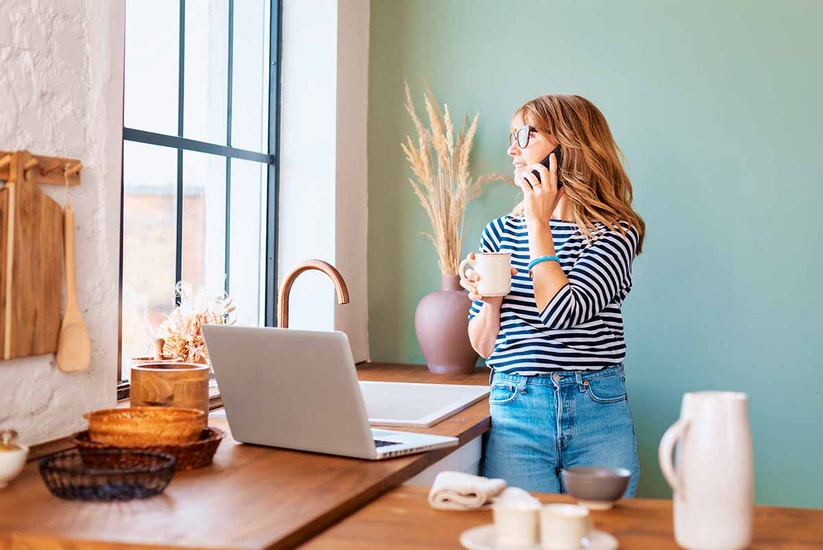 Dame står på kjøkkenet med en kopp i hånda og telefonen mot øret, til illustrasjon for svindel