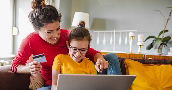 Mor og barn sitter på sofa og ser på datamaskin med bankkort i handa, til illustrasjon for barn og penger