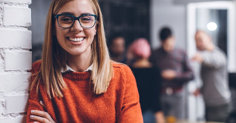 Kvinne med orange genser og briller smiler i kontorlokale, til illustrasjon for bli bedriftskunde