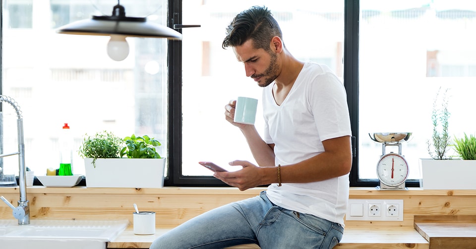 Mann sitter på kjøkkenbenk med kaffe og mobil i hendene